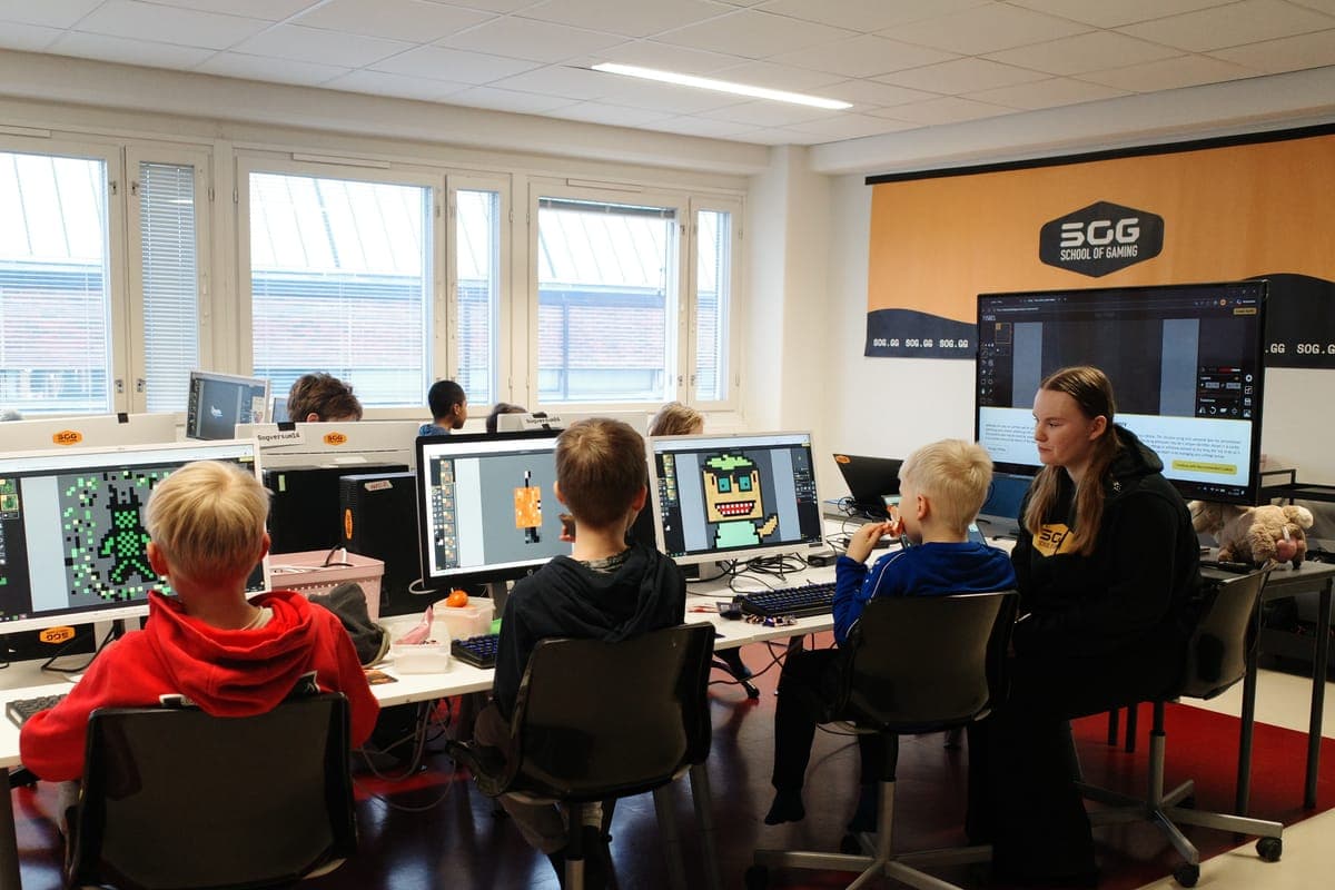 Des enfants devant des ordinateurs dans un club School of Gaming, vus de dos, avec leur animateur assis à côté d’eux